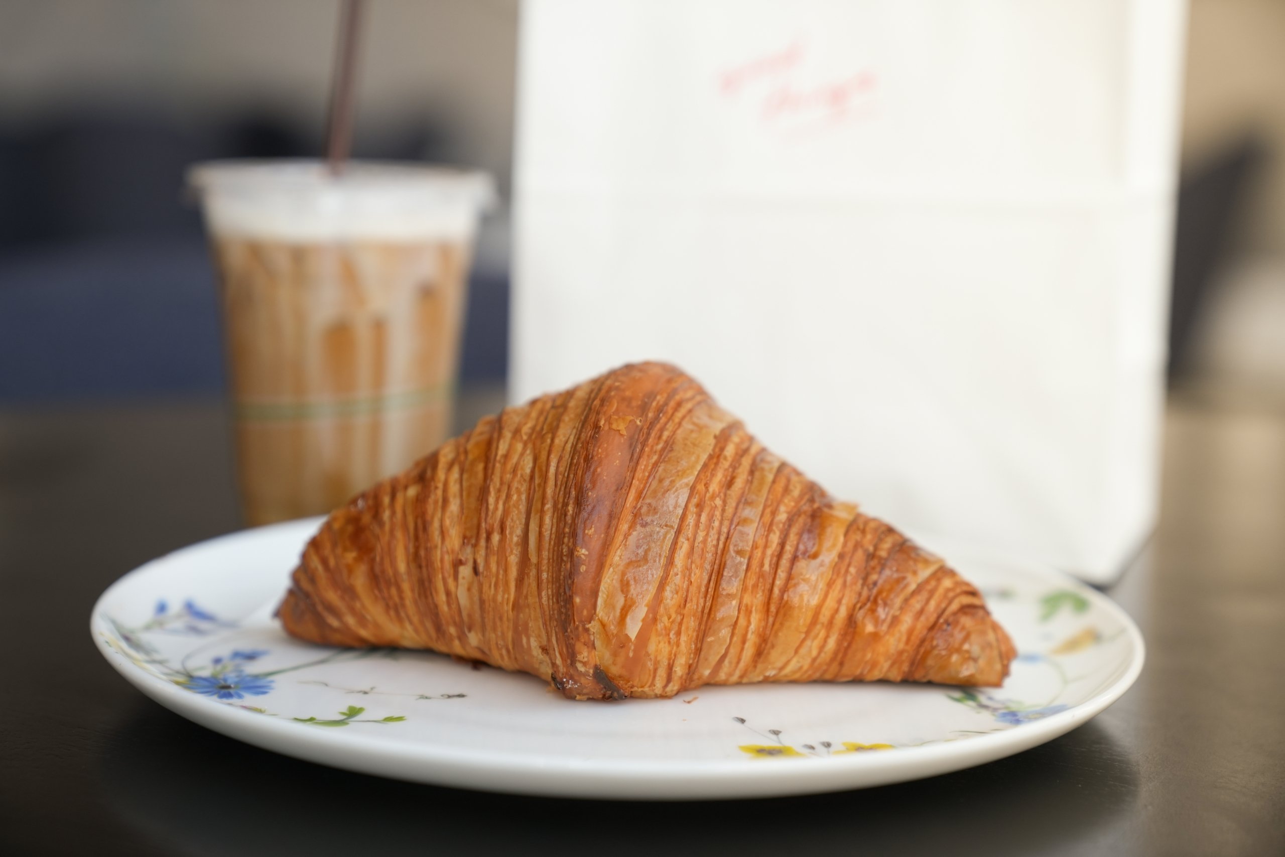 A butter croissant sits on a plate in front of a coffee drink